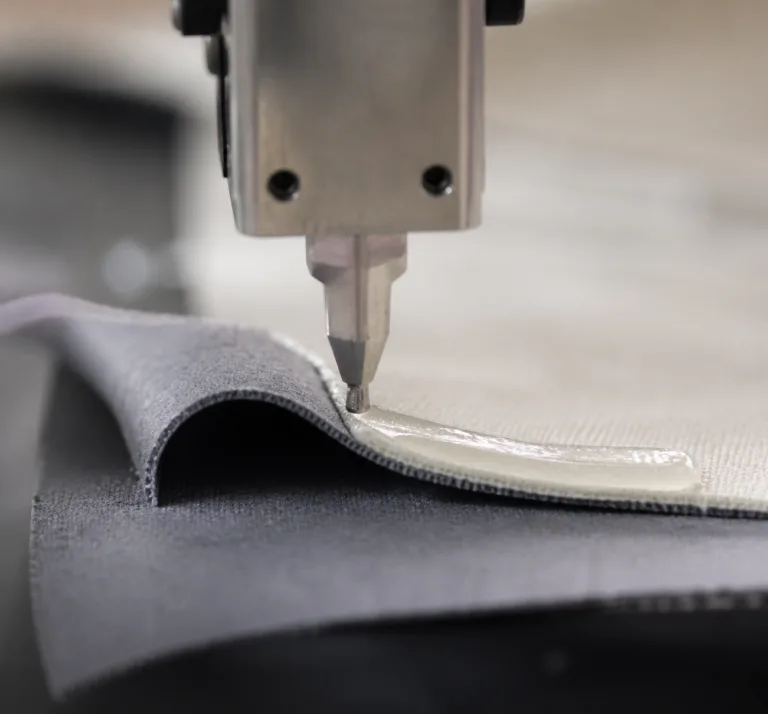 Material 8 A realistic close-up showing fabric layers being bonded with adhesive in an industrial manufacturing setting, with visible adhesive application and professional product photography styling.
