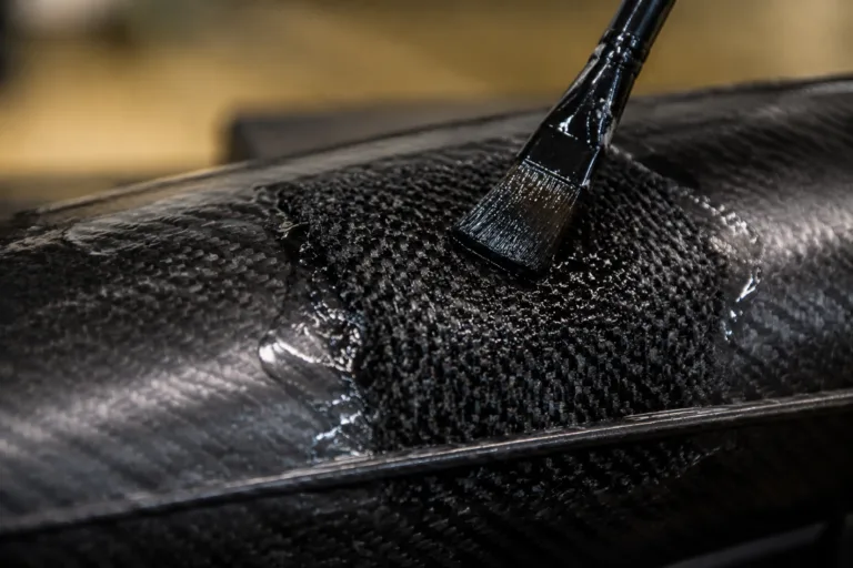 Carbon Fiber Adhesive 13 A realistic close-up scene showing carbon fiber repair adhesive applied to a localized patch for restoration and reinforcement, with detailed woven carbon fiber texture and a professional workshop setting.